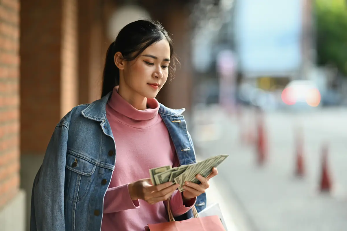 A woman standing outdoors counts a stack of cash while holding a shopping bag.