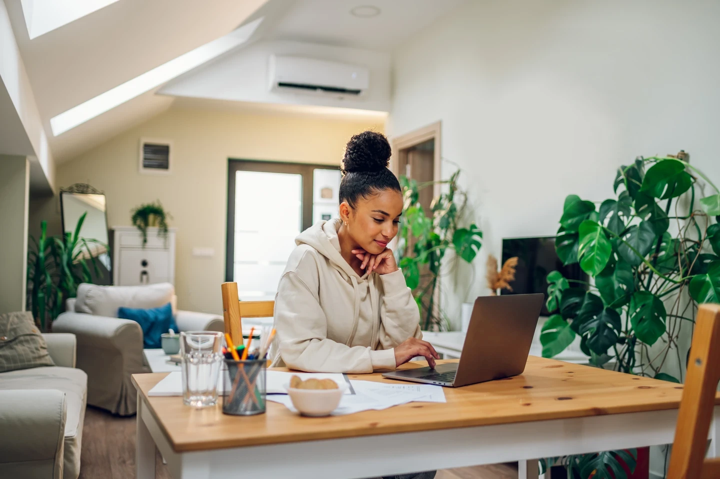 A person sits at a wooden table in a cozy, well-lit room, working on a laptop surrounded by papers and plants.