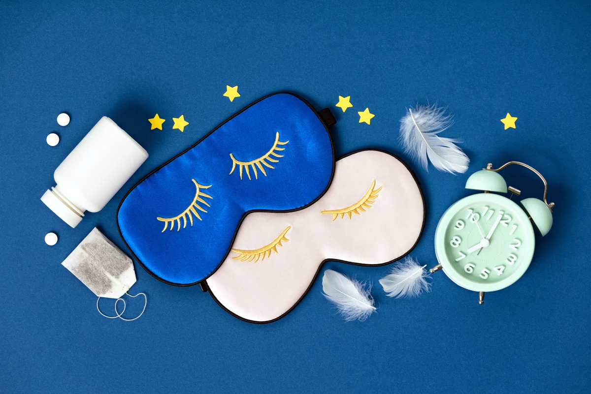 Sleep masks, alarm clock, and pills on blue background.