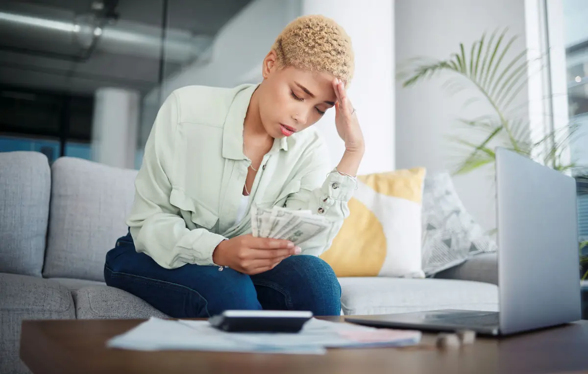 A person sitting on a couch looking stressed while holding cash, with bills and a calculator on the table in front.