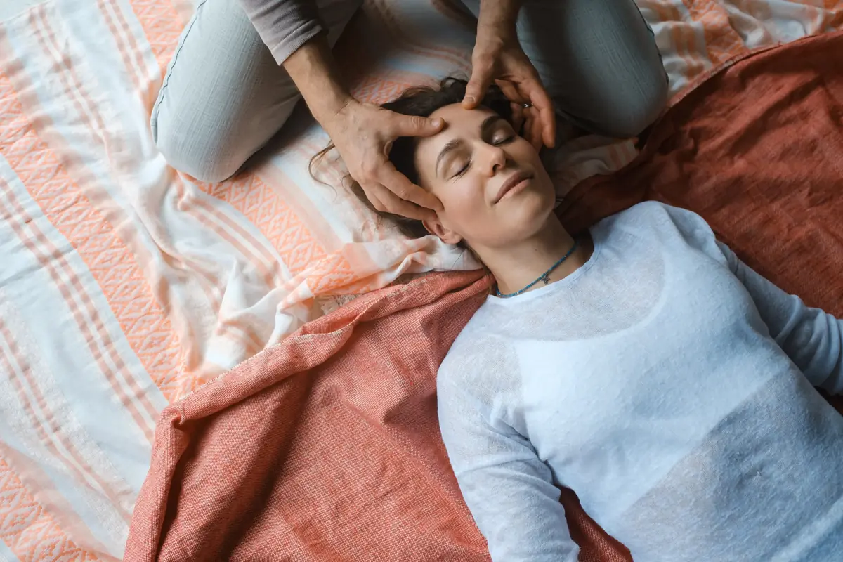 A person lying down with closed eyes, receiving a gentle forehead massage from hands positioned above them on a soft, patterned fabric.