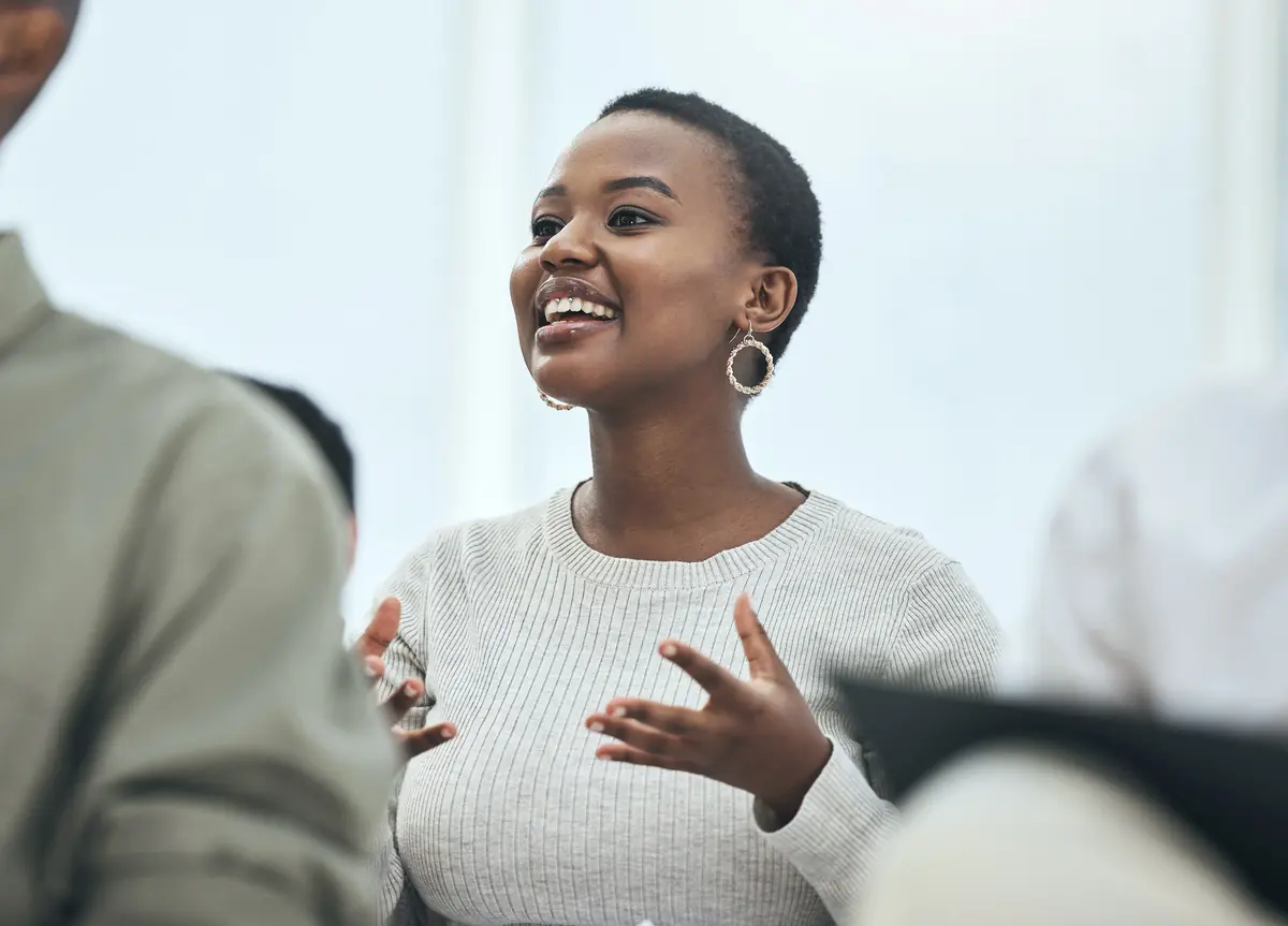A woman speaking passionately in a group setting, gesturing with her hands, wearing a light sweater and hoop earrings.