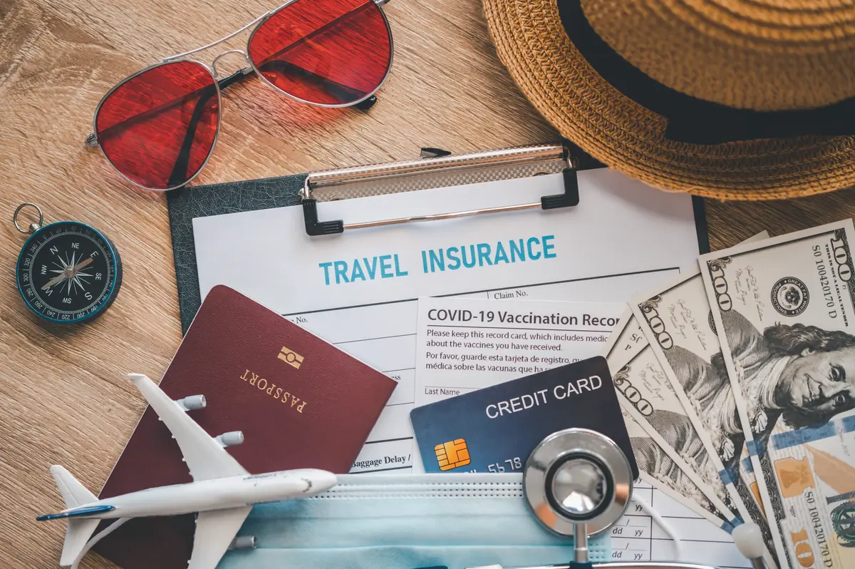 Travel essentials including a travel insurance form, passport, miniature airplane, red sunglasses, compass, hat, vaccination record, credit card, and U.S. dollar bills arranged on a table.