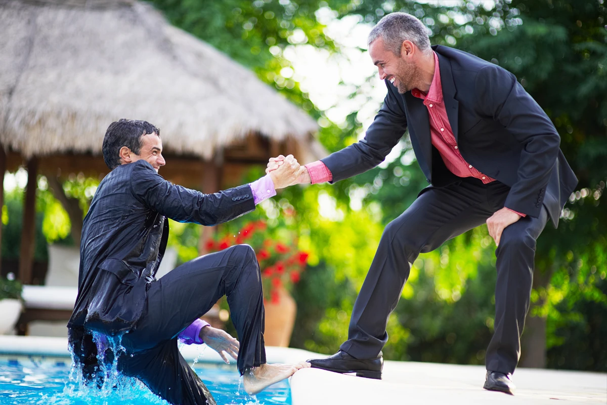 A man in a wet suit is being playfully pulled out of a swimming pool by another man in a dry suit, both smiling and surrounded by a tropical outdoor setting.