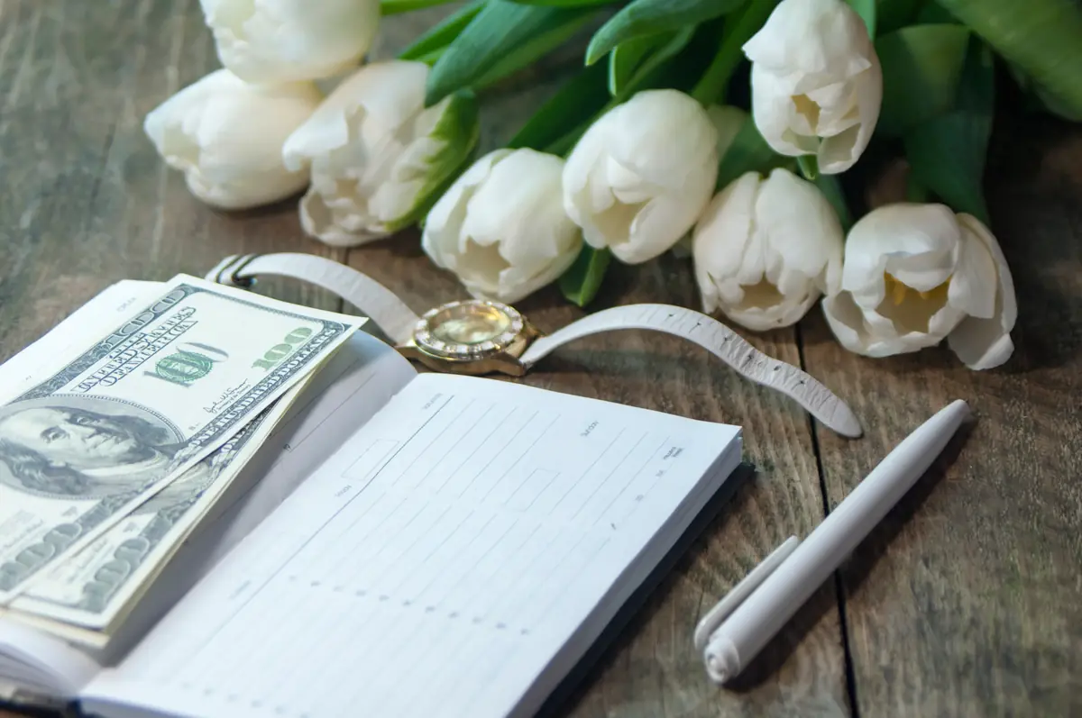 Open planner with blank pages, a stack of hundred-dollar bills, a gold wristwatch, and a bouquet of white tulips on a wooden table.