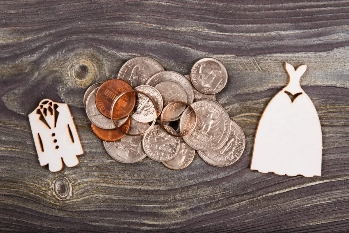 Wooden cutouts of a tuxedo and wedding dress flank a pile of coins, symbolizing wedding costs.