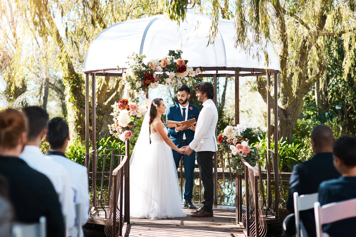 Bride and groom hold hands in an outdoor wedding ceremony under a floral-decorated gazebo, surrounded by lush greenery and witnessed by seated guests.