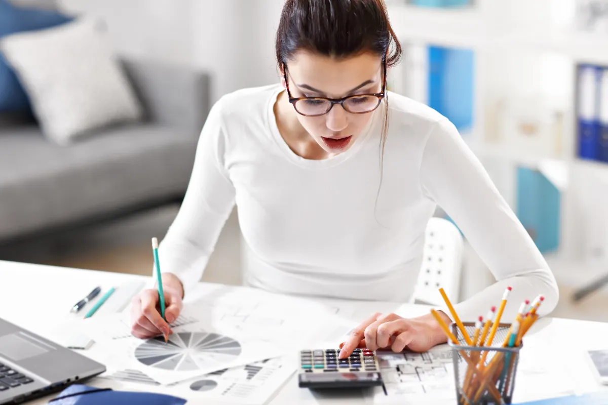 Young woman wearing glasses is focused on budgeting at a desk with a calculator, pie chart, and numerous papers.