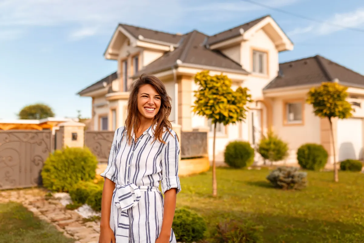 A smiling woman stands in front of a modern two-story house with a manicured lawn and small trees.
