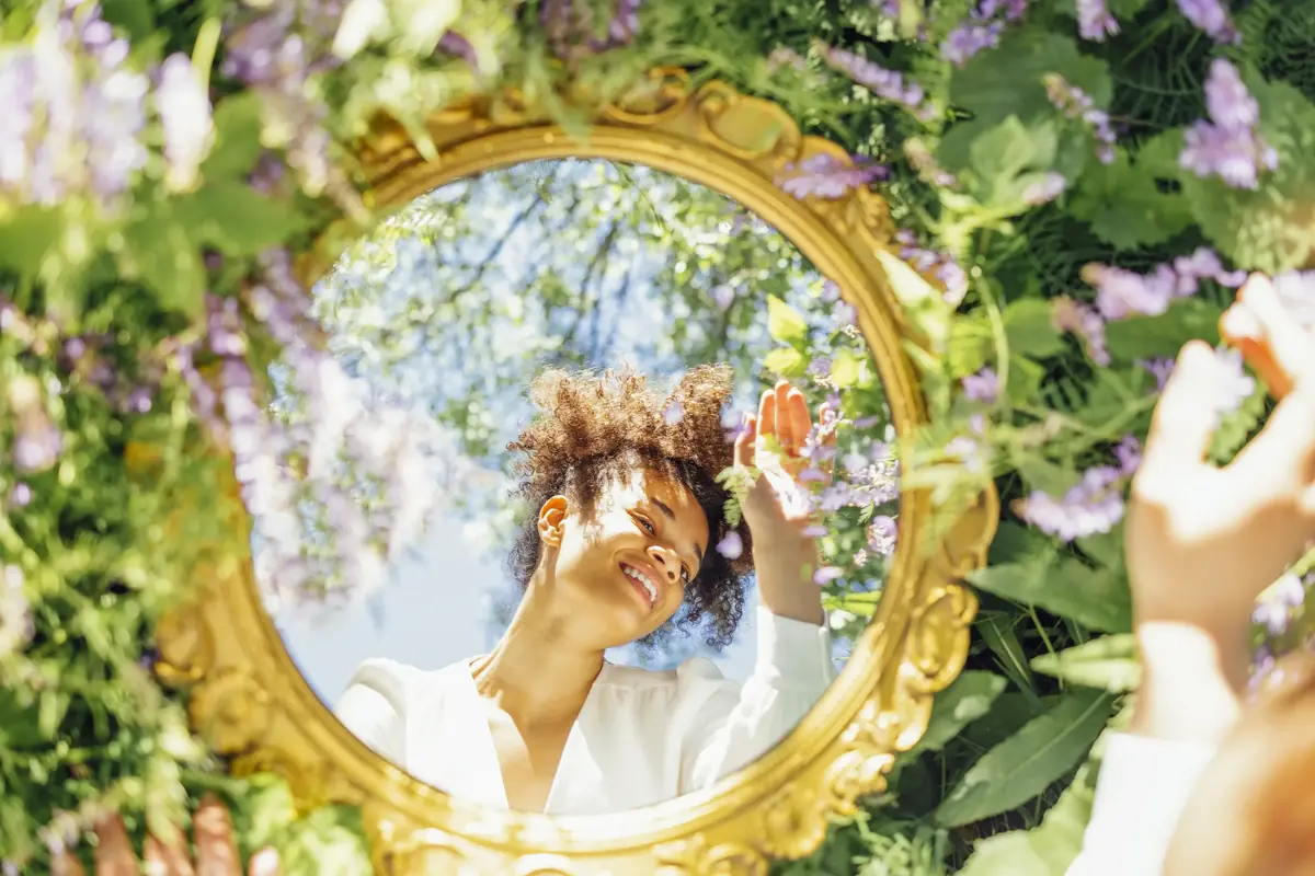 A woman with curly hair gazes introspectively at her reflection in an ornate circular mirror surrounded by vibrant green leaves and purple flowers, under a clear blue sky.
