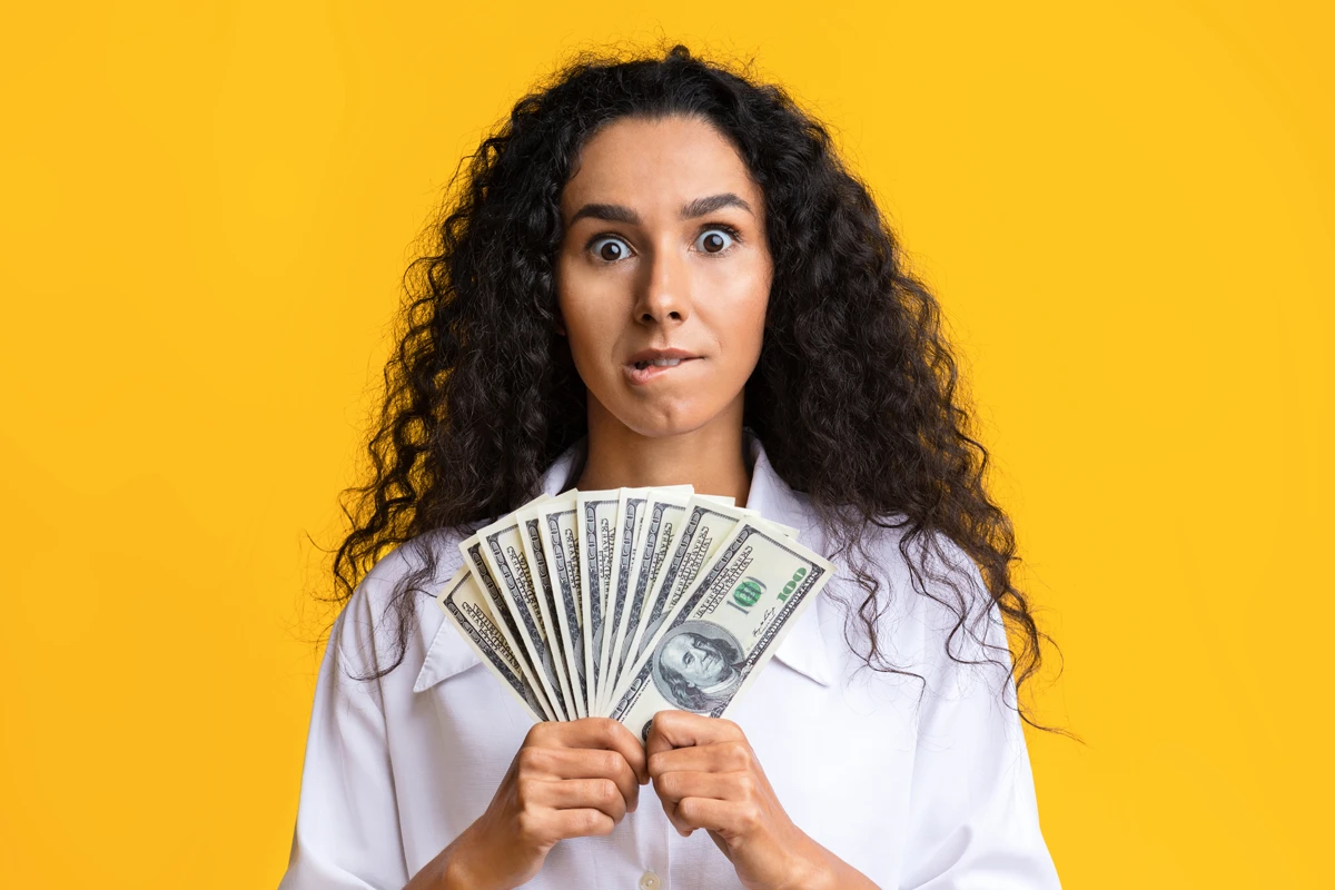 Surprised woman holding dollar bills on yellow background.