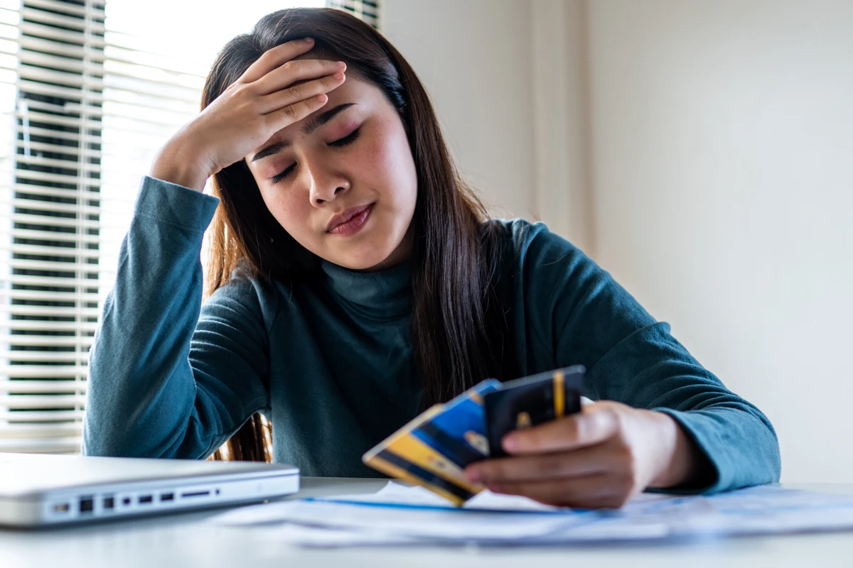 A woman sitting at a table, holding her forehead with one hand and credit cards with the other, looking distressed, with bills and a laptop in front of her.