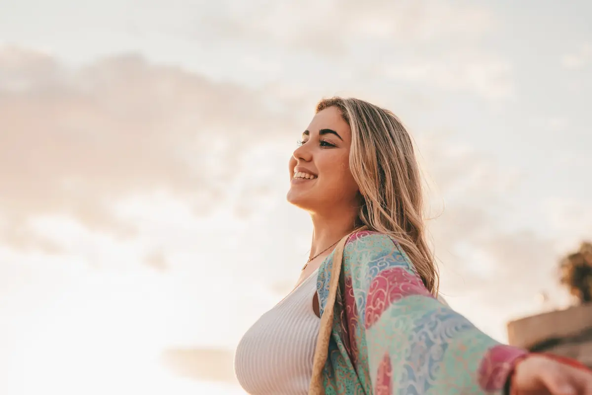 A smiling woman stands outdoors with her arms outstretched, wearing a colorful shawl under a vibrant sky.
