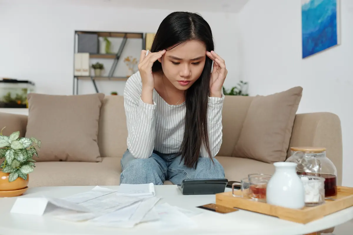 Young woman sitting on a couch in a living room, focused on a calculator with scattered bills and documents on the table in front of her.