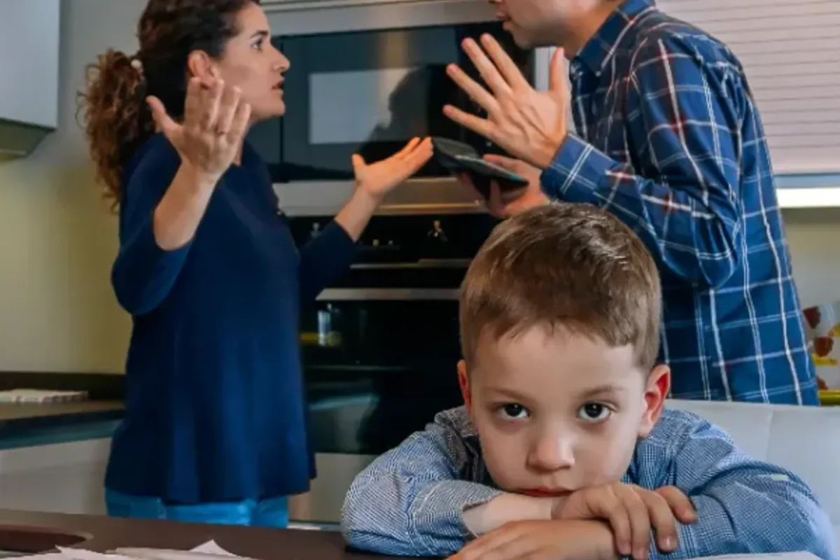 A young boy looking sad with his head resting on his arms at a kitchen table cluttered with papers, while a man and woman appear to be arguing in the background.