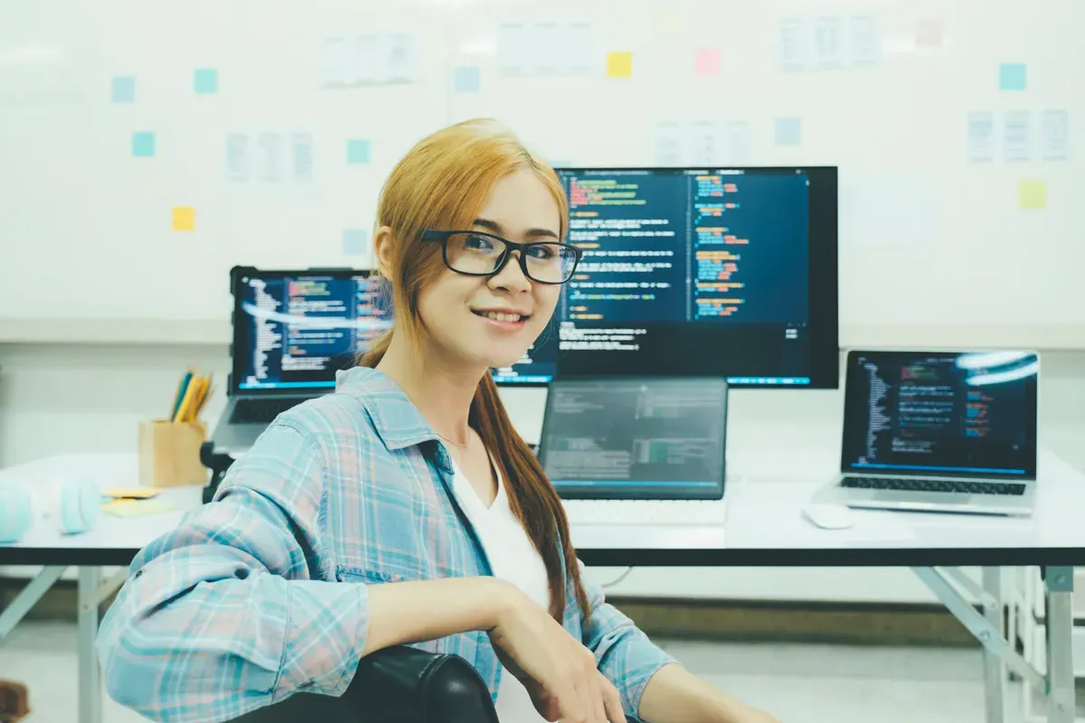 A young woman with glasses smiles while seated in an office filled with monitors displaying code. The workspace is organized and collaborative.
