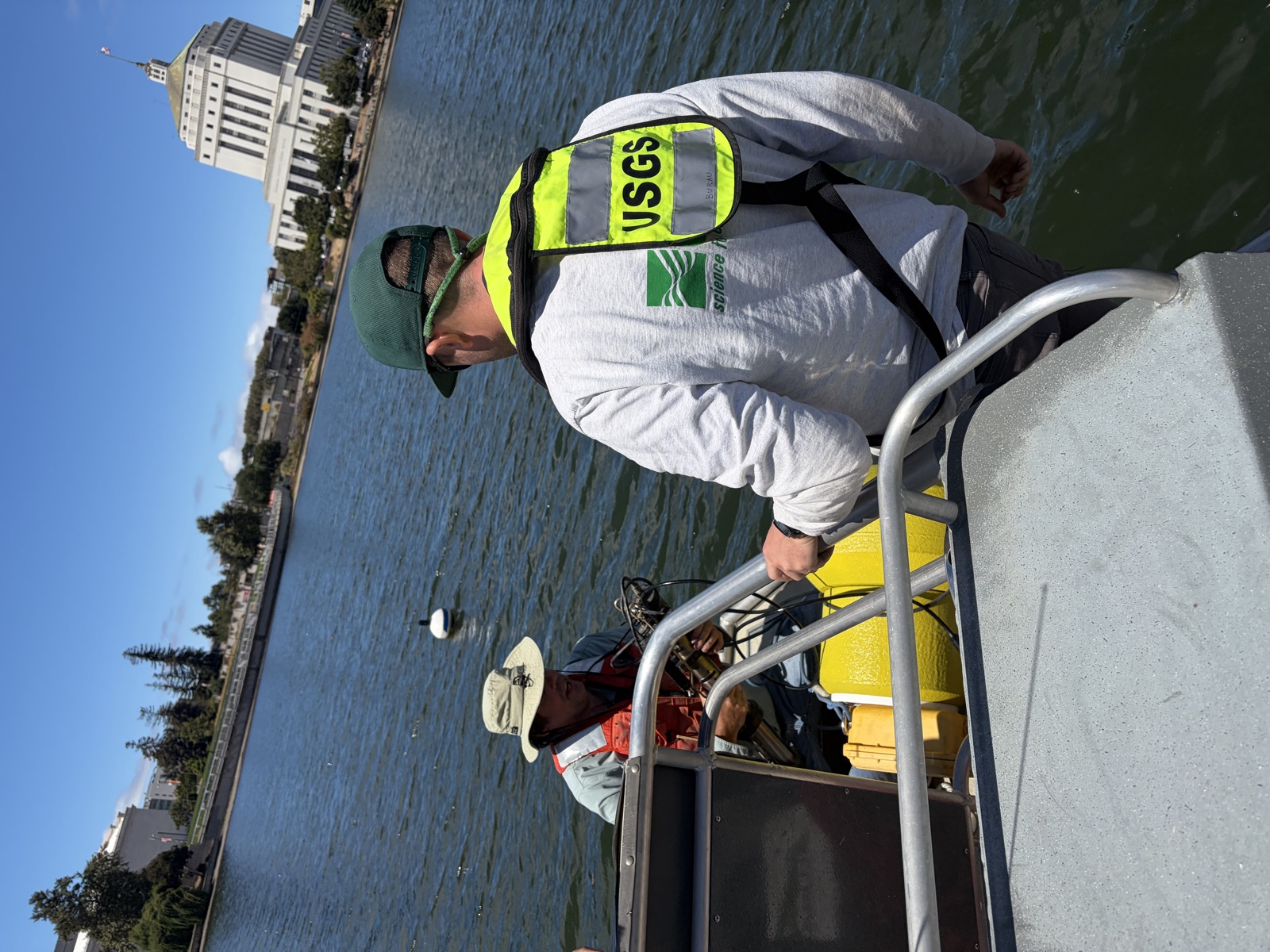 USGS crew member in a safety vest and a technician in an orange life jacket preparing sensor hardware on the deck of a workboat at Lake Merritt