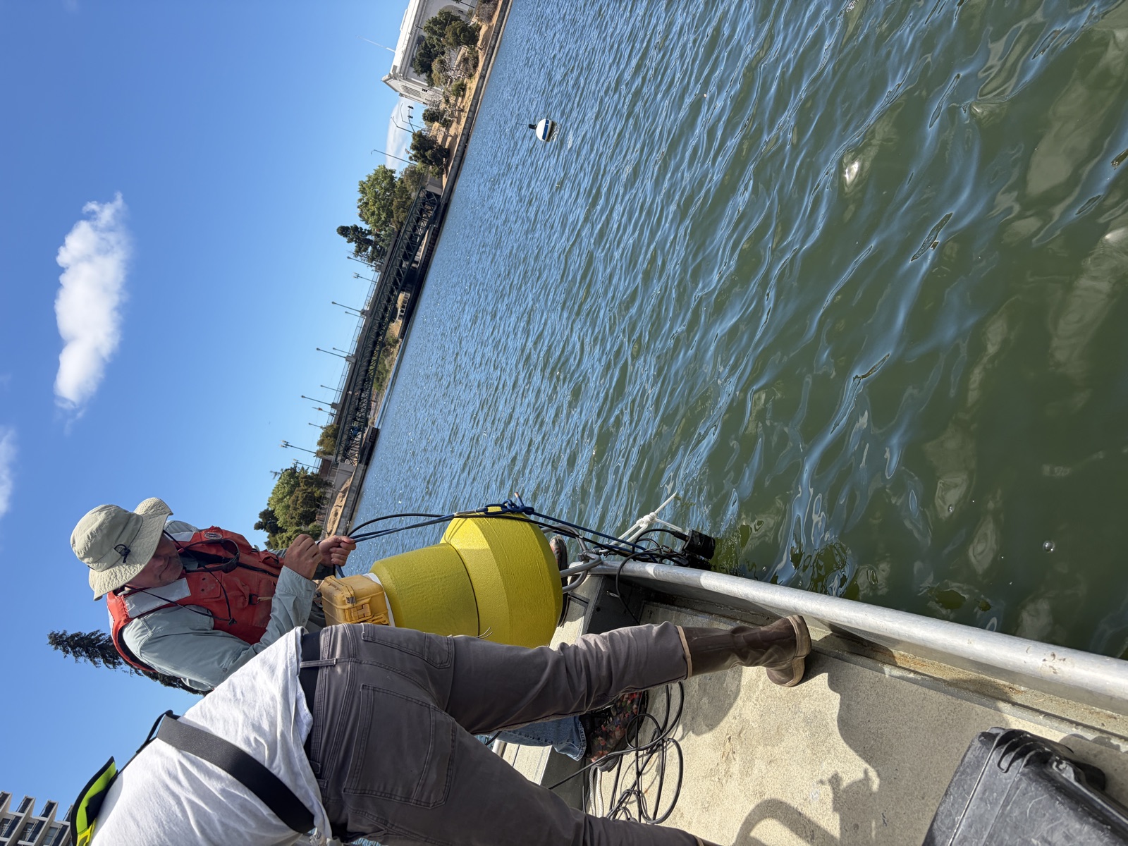 Technicians lowering the yellow monitoring buoy with attached sensor cables over the side of the boat into Lake Merritt