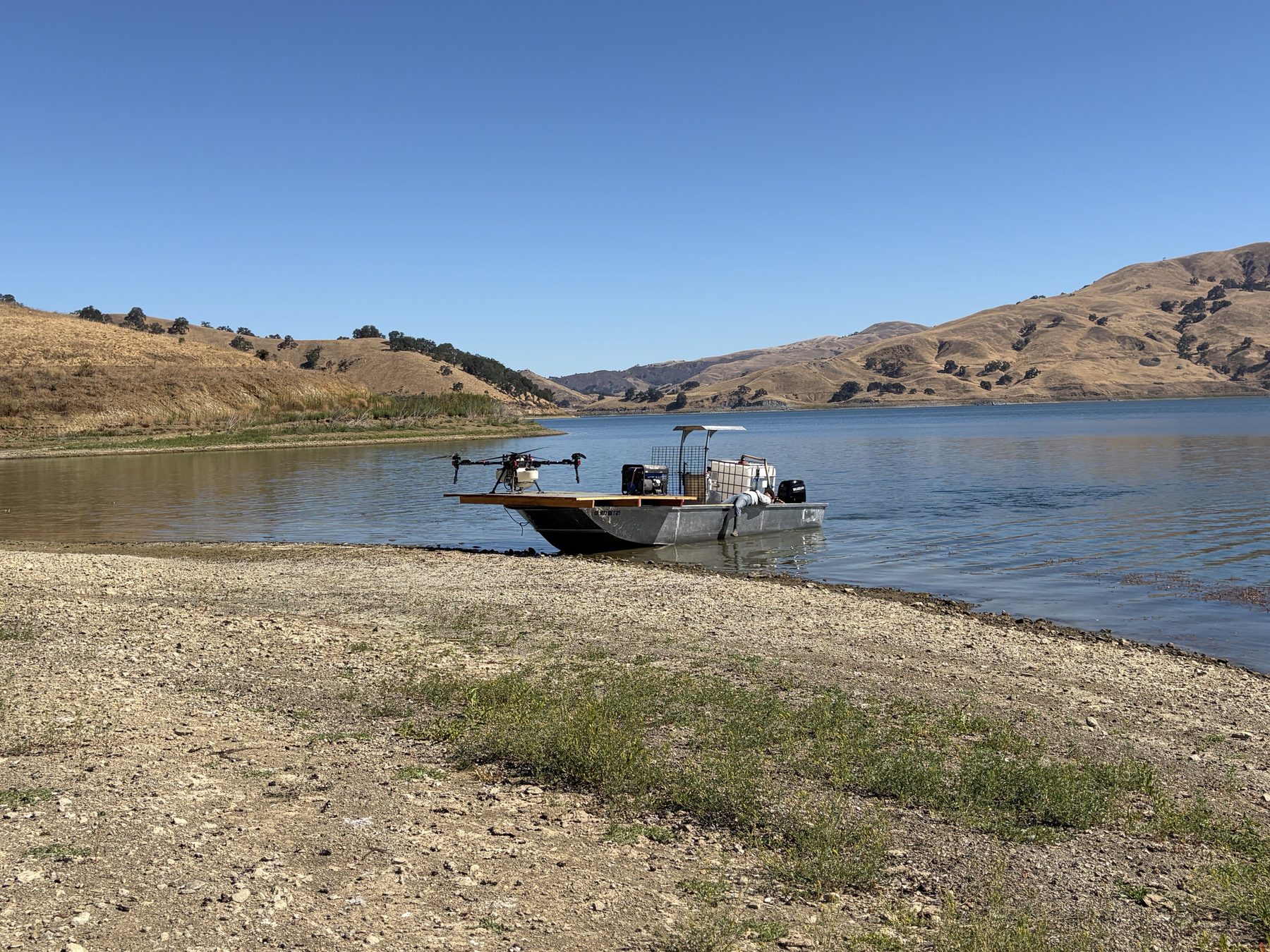 A flat deck aluminum barge carrying a spray drone, beached on the shoreline of Calaveras Reservoir with golden hills behind it