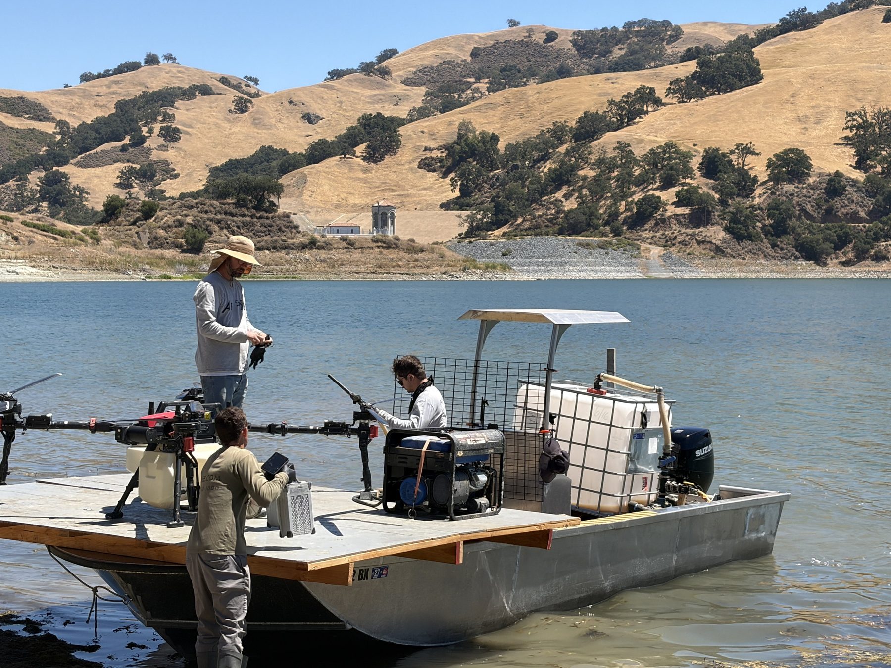 Three crew members rigging a spray drone on the deck of a barge at Calaveras Reservoir, with the dam intake tower in the background