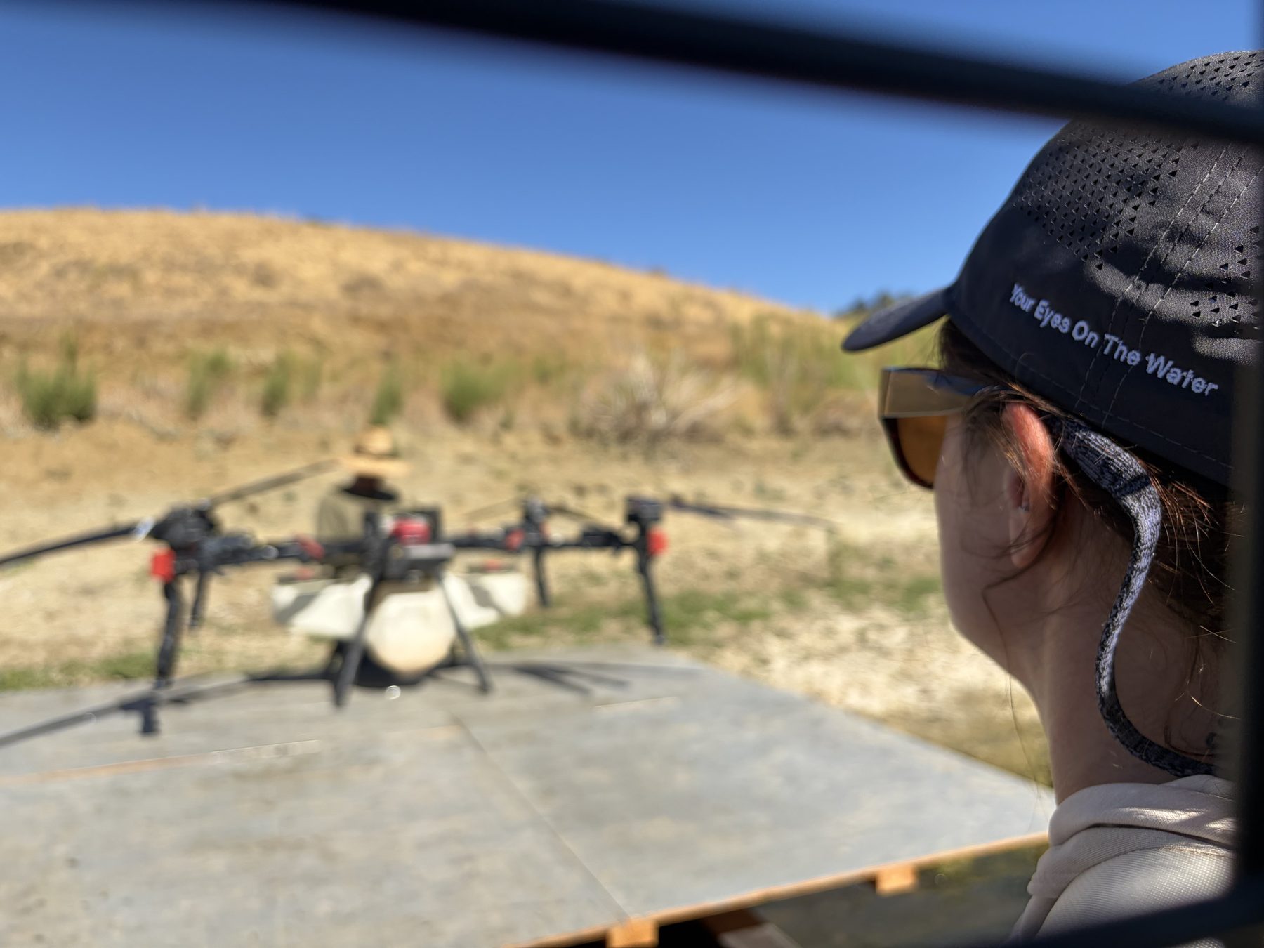 Over the shoulder view of a person in a LakeTech cap watching a spray drone on its launch pad above Calaveras Reservoir