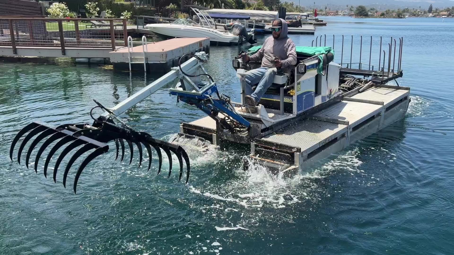 An amphibious Truxor lake management machine swinging a wide raking attachment over the surface of a residential lagoon, with private docks and moored boats behind it
