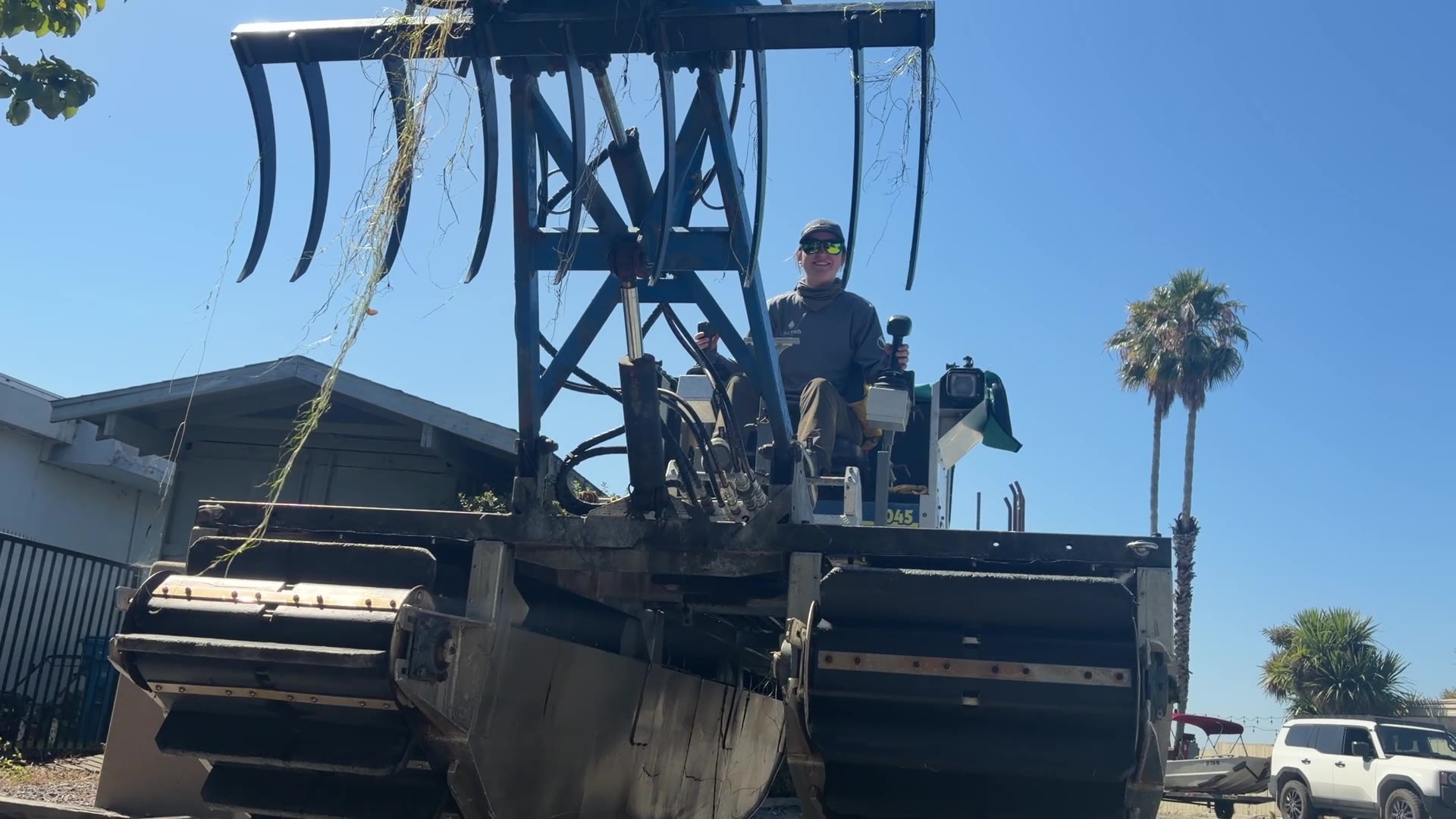 Low angle view of a Truxor sitting on dry ground, showing the wide tracked pontoons it runs on and a raised rake attachment still carrying aquatic vegetation