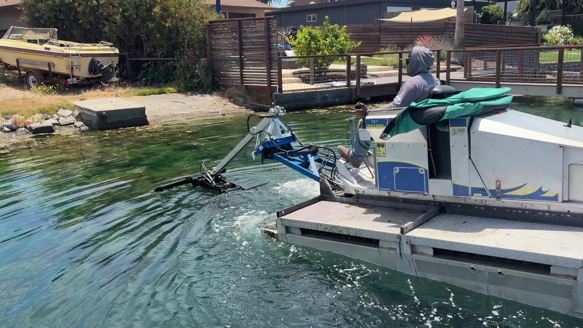 A Truxor working a shallow lagoon edge alongside a private dock, with its cutting attachment lowered below the waterline