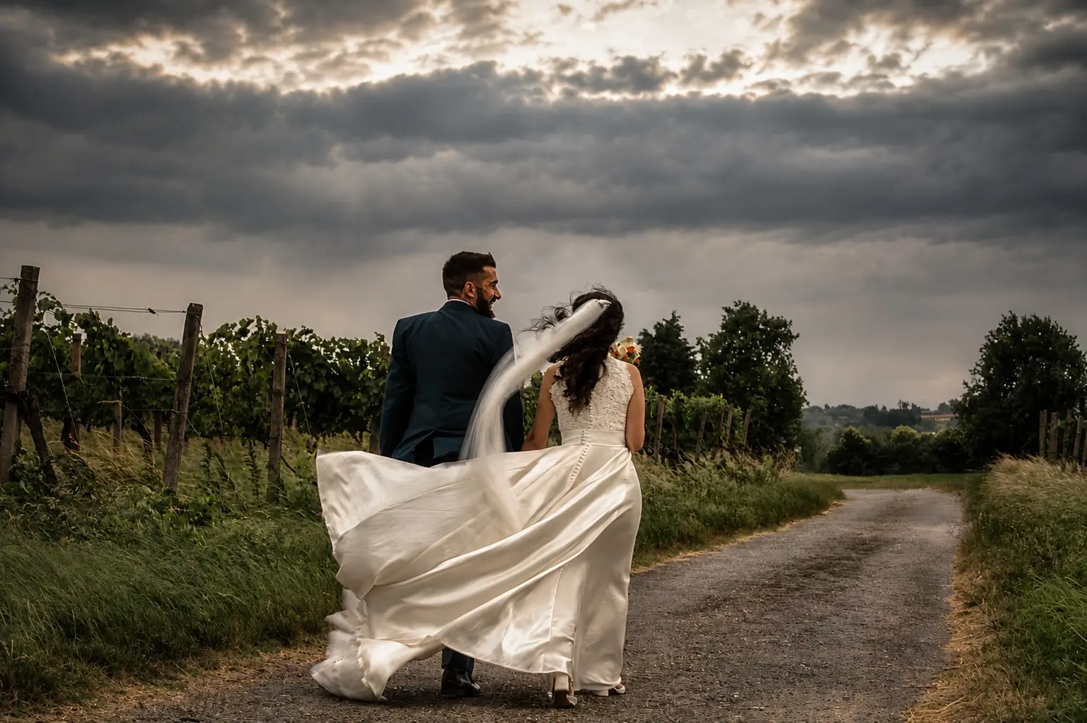 Sposi che camminano su una strada di campagna tra i vigneti sotto un cielo di nuvole