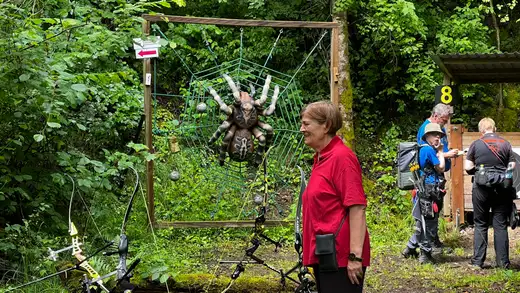 Teilnehmer am Bogenschieß-Wettkampf blicken auf ein großes Spinnennetz-Ziel mit einer Spinne in der Mitte. Ein Schütze im Vordergrund steht neben ein.