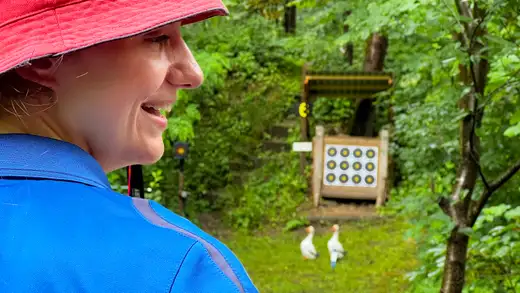 Teilnehmerin mit roter Kappe und blauem Polohemd lächelt, im Hintergrund eine Zielscheibe und zwei Gänsefiguren.