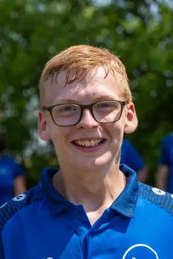 Junger Schütze mit Brille und roten Haaren lächelt in die Kamera, im Hintergrund unscharfe Personen in blauen Trikots.
