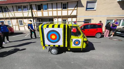Gelber Kleintransporter mit Zielscheiben und schwarz-gelben Streifen auf einer Straße vor Fachwerkhäusern.