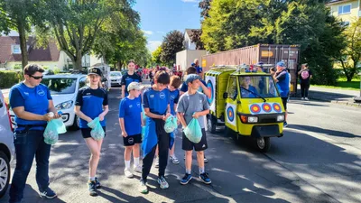 Teilnehmer in blauen Vereinsshirts gehen mit Beuteln neben dem geschmückten gelben Kleintransporter.