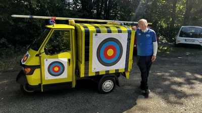 Gelber Kleintransporter mit großer Zielscheibe an der Seite und überlangem Pfeil auf dem Dach, daneben ein Vereinsmitglied.