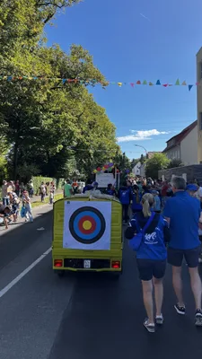 Der gelbe Wagen fährt zwischen Zuschauern durch eine mit bunten Wimpeln geschmückte Straße; Mitglieder gehen daneben.