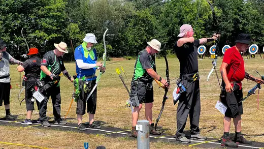 Mehrere Bogenschützen stehen in einer Reihe und bereiten sich auf den Schuss vor. Sie tragen unterschiedliche Kleidung und Hüte, einige haben Pfeile.