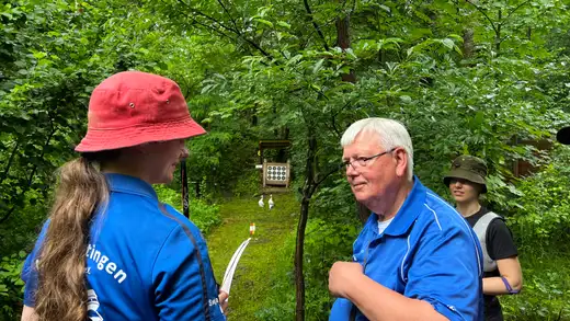 Zwei Bogenschützen unterhalten sich am Feldbogenparcours. Im Hintergrund ist eine Zielscheibe zu sehen.