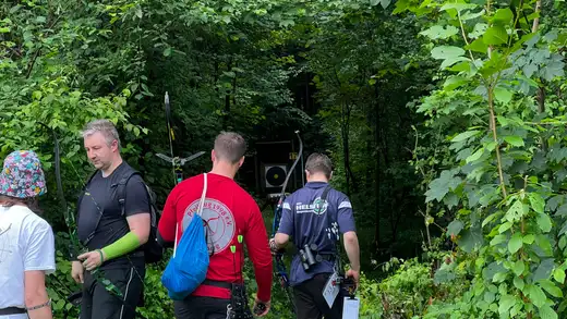 Mehrere Bogenschützen gehen durch einen Waldweg zu einer Zielscheibe. Ein Schütze im Vordergrund trägt einen grünen Armschutz.