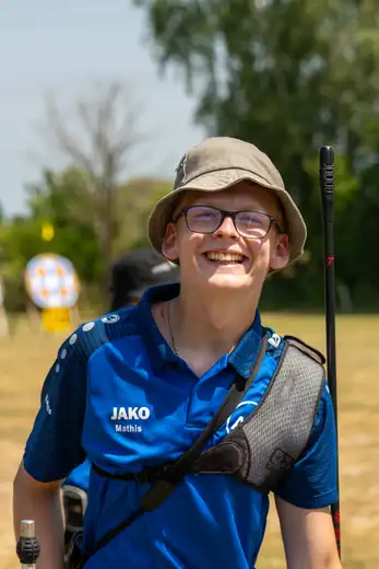Ein junger Schütze mit Hut und Brille lächelt in die Kamera. Er trägt ein blaues JAKO-Shirt und eine Schießweste. Im Hintergrund ist eine Zielscheibe.