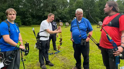 Teilnehmer mit Bögen und Pfeilen stehen auf einer Wiese vor Zielscheiben. Ein Schütze erklärt einem anderen die Handhabung eines Bogens.