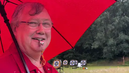 Ein Mann mit Brille und rotem Hemd hält eine rote Regenschirm-Stange und eine Pfeife im Mund. Im Hintergrund sind mehrere Zielscheiben aufgestellt.