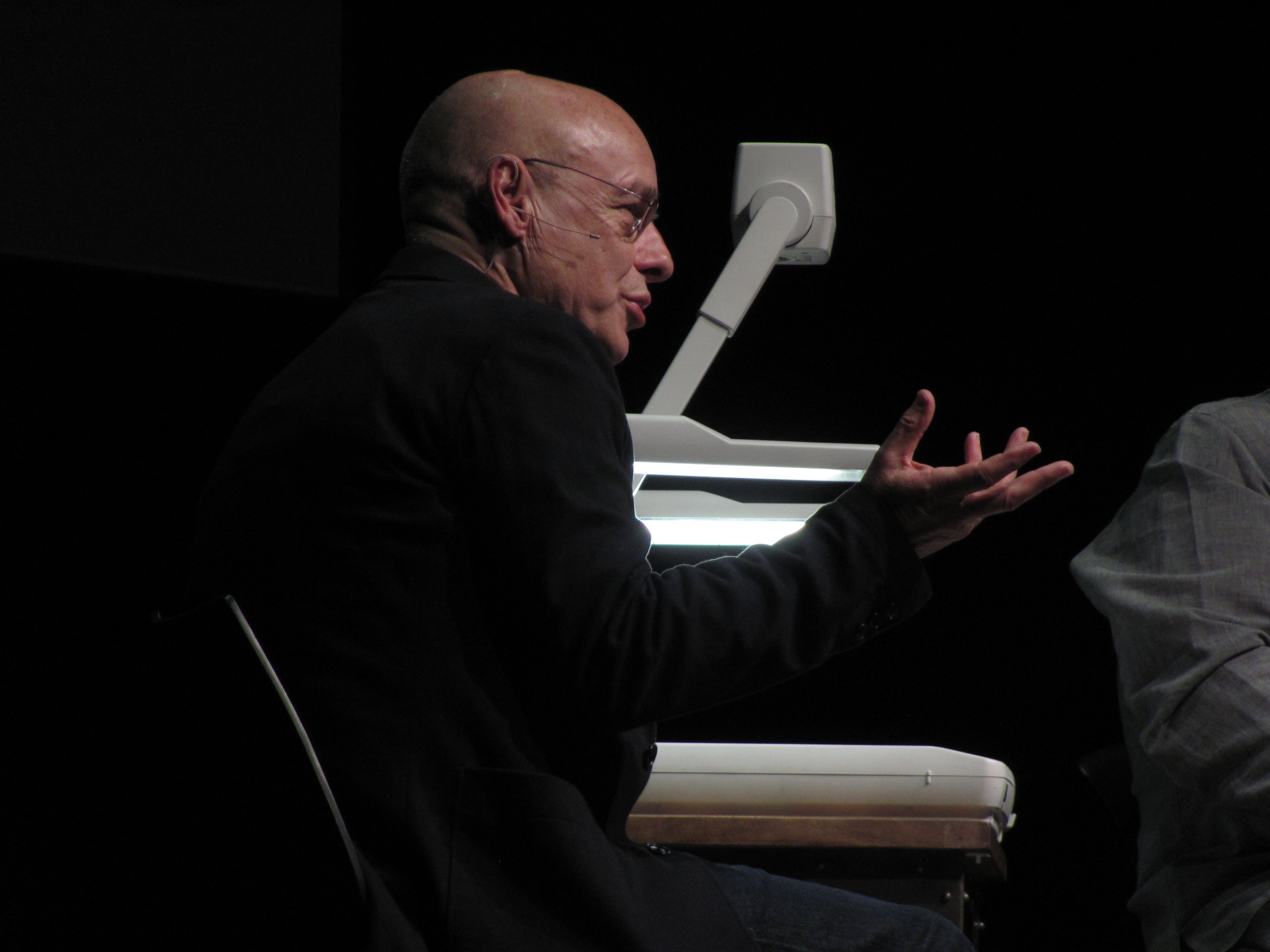 Brian Eno sitzt auf einer Bühne und spricht mit einer Handbewegung in Richtung Publikum.