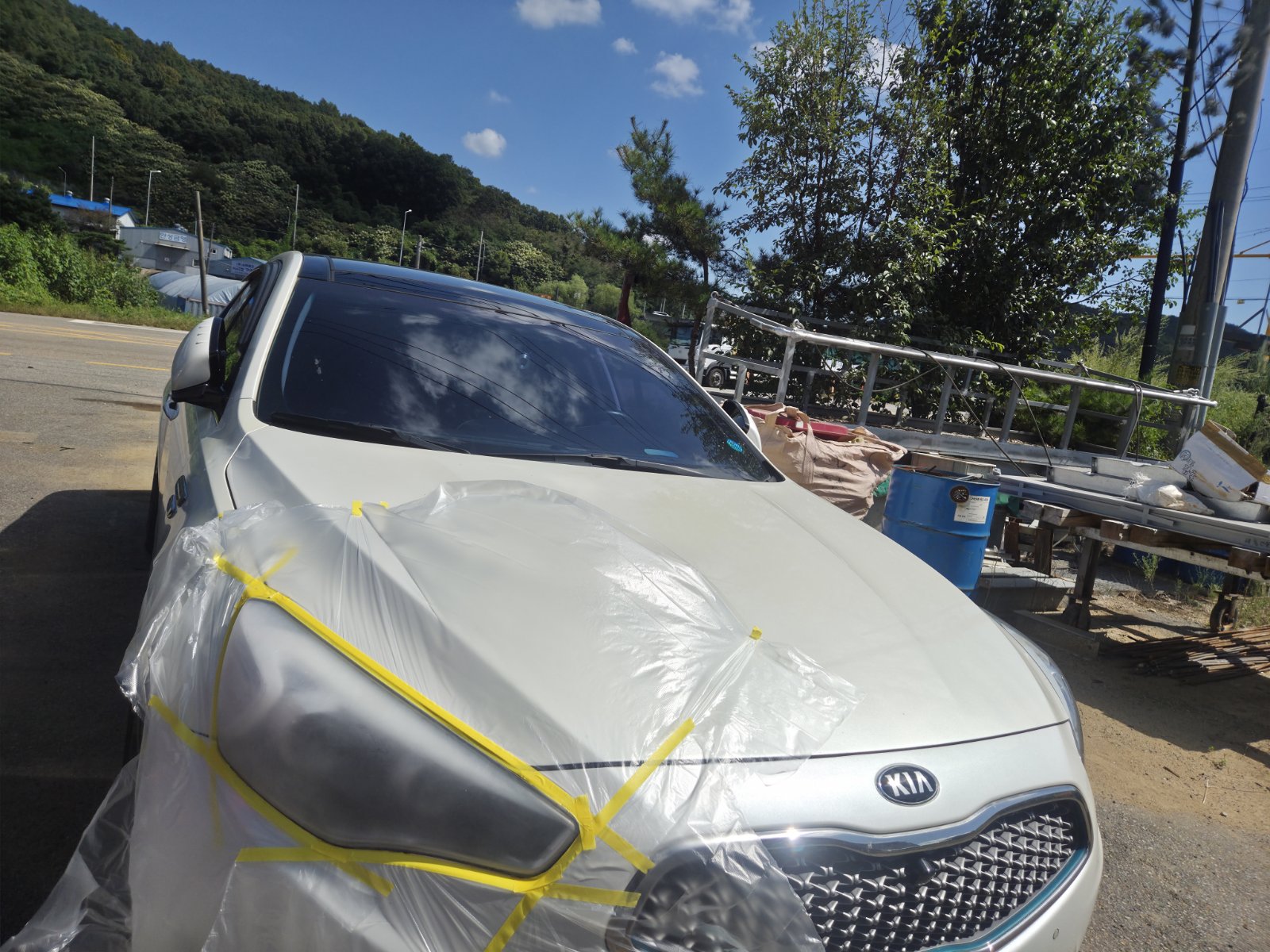 보호 비닐과 노란 마스킹 테이프로 헤드라이트와 보닛을 가린 채 작업 중인 은색 KIA 차량 전면 사진