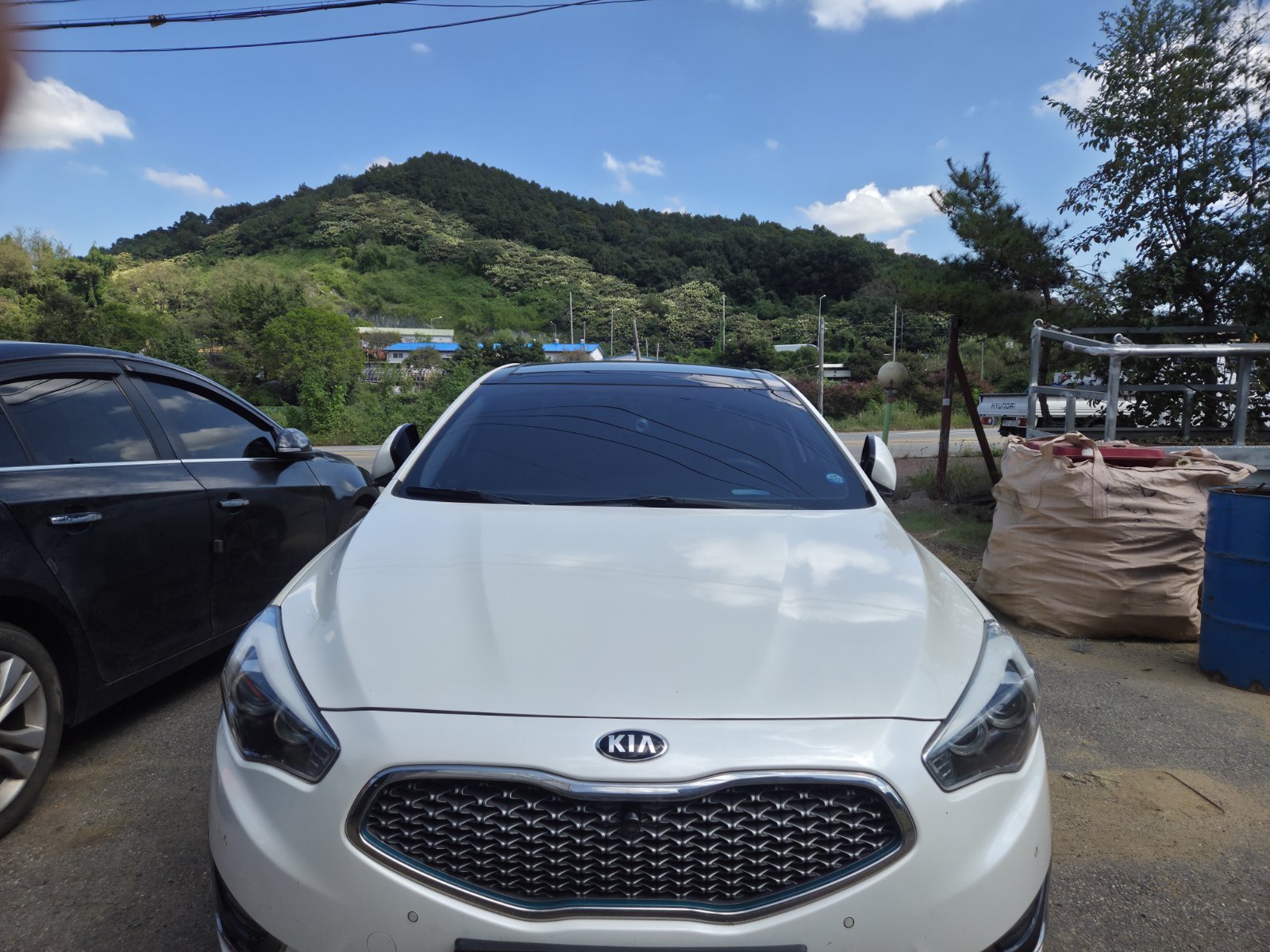 흰색 KIA 승용차 전면 사진으로, 작업장 옆 야외 주차장에 세워져 있고 배경에 녹색 언덕과 파란 하늘이 보인다.