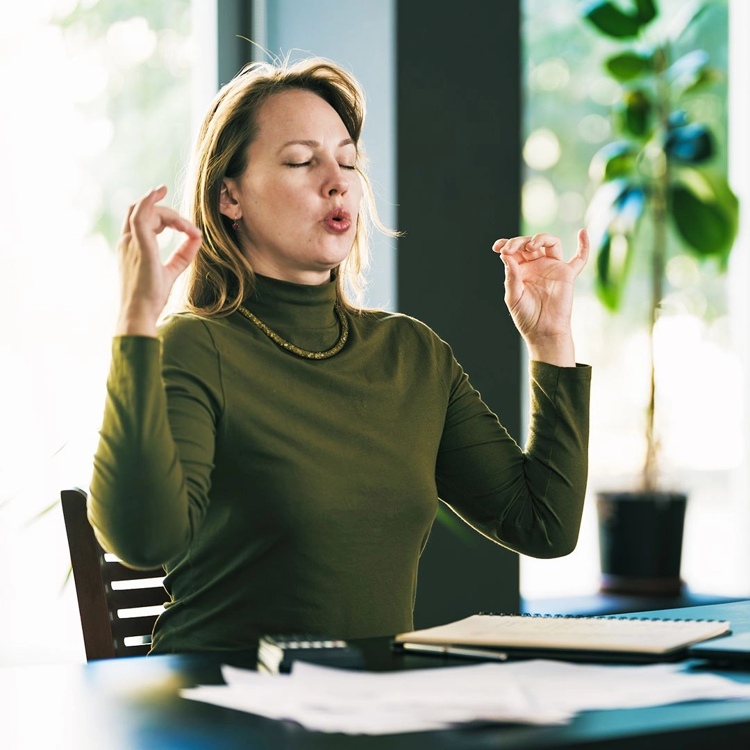 Blonde woman wearing an olive-green turtleneck top is sitting at a desk with a calculator and papers. She has her eyes closed and hands up in a calming position while she is deeply breathing for relaxation.