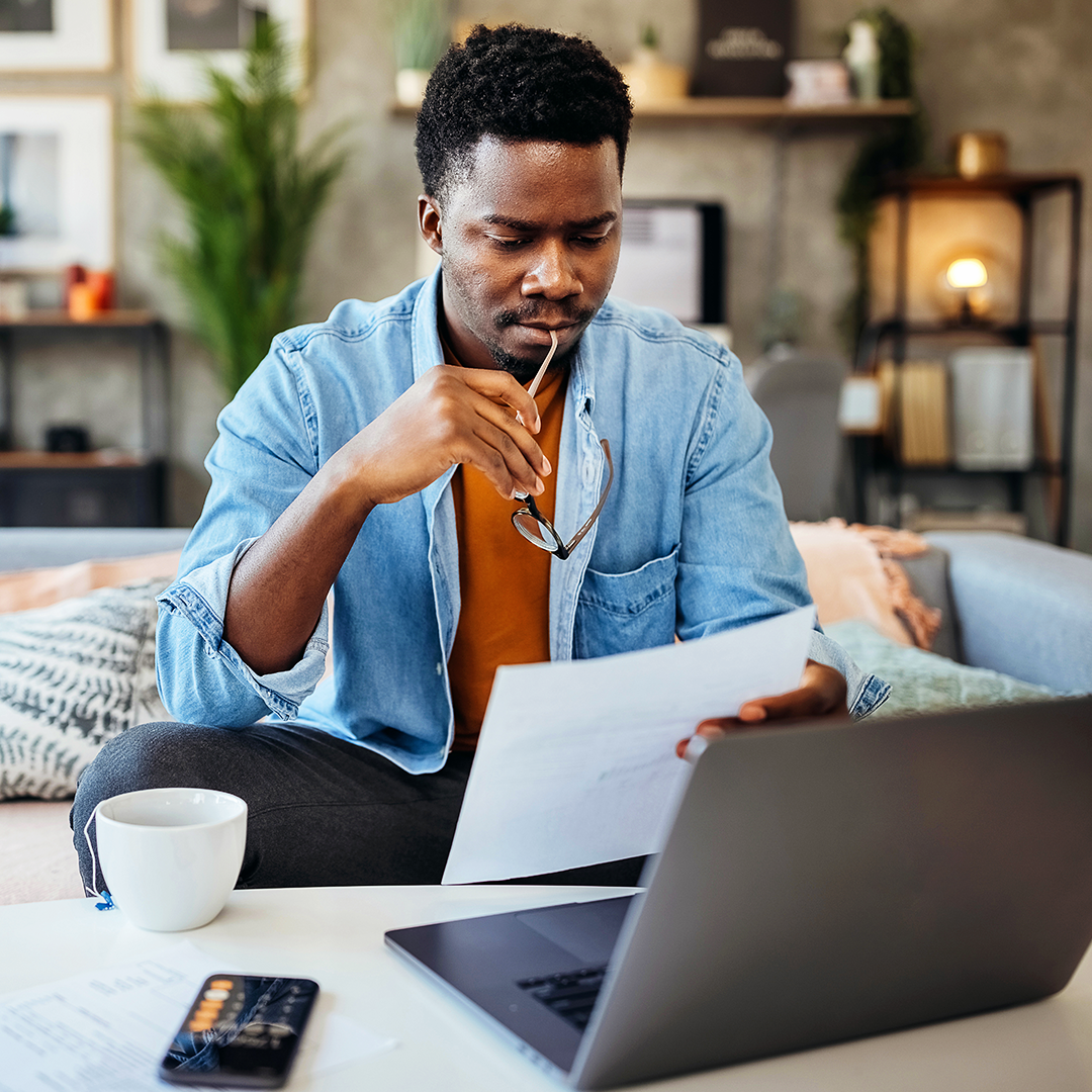 Image of a man holding eyeglasses and a piece of paper, contemplating in front of a laptop, calculator and cup of tea.