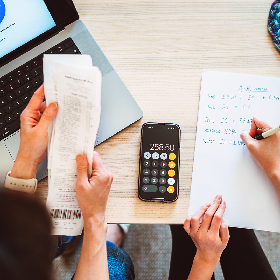 Image of two peoples’ hands working a monthly budget in front of a laptop, calculator, receipts, and handwritten budget.