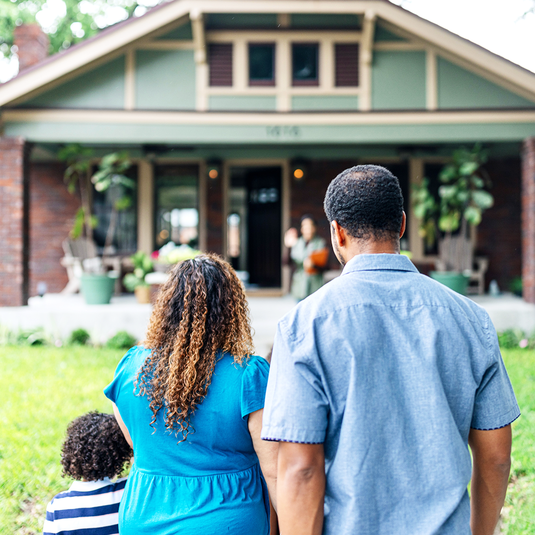 Image of the back of a family (man, woman, and young child) walking up to a green and red brick home to meet their realtor.