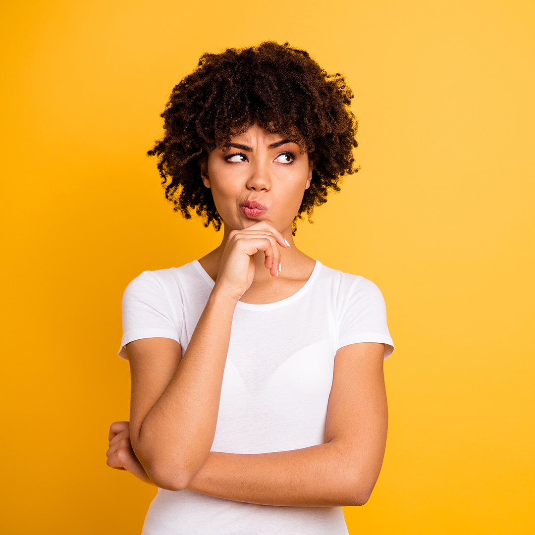 Image of a woman with short curly hair wearing a white t-shirt in front of a yellow background. Her facial expression and arm placement show she is thinking about something.